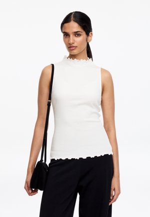 Femme aux cheveux foncés attachés, portant un haut blanc sans manches à bords froncés et un pantalon noir, tenant un sac à bandoulière noir.