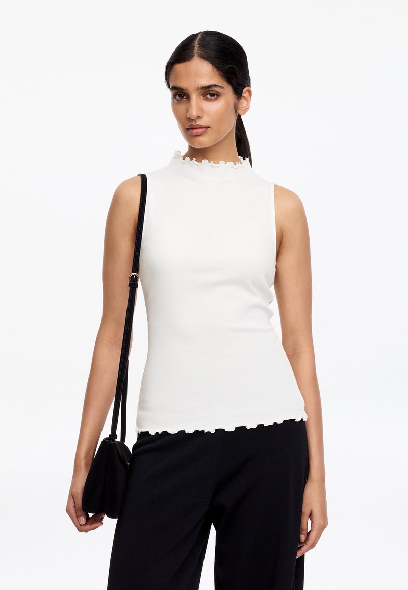 Femme aux cheveux foncés attachés, portant un haut blanc sans manches à bords froncés et un pantalon noir, tenant un sac à bandoulière noir.