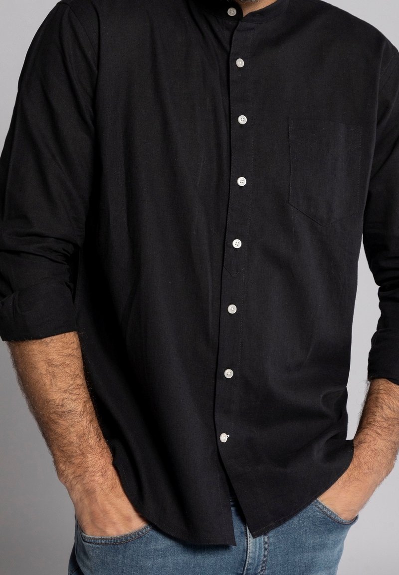 Uomo che indossa una camicia nera con bottoni e maniche arrotolate, jeans blu, mani casualmente nelle tasche, sfondo grigio.