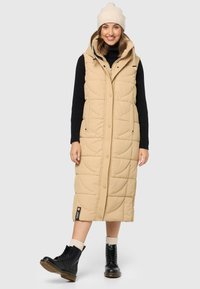 Beige dlhý puffový vest s kapucňou, prešívaná textúra, bočné vrecká, skombinovaný s čiernym rolákom, krémovou baretou a čiernymi šnurovacími topánkami.