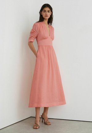 Nő korallrózsaszín midi ruhát visel V-nyakkal, összegyűrt derékkal, könyékig érő ujjakkal és világosbarna magas sarkú szandállal, egy fehér sarkú fal előtt állva.