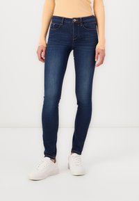 Skinny jeans in dark blue denim, featuring a high waistband, front pockets, and subtle stitching details, paired with white sneakers.