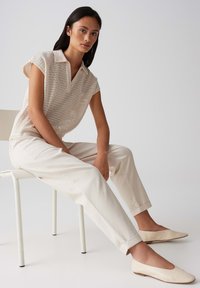 Haut en tricot beige avec col, associé à un pantalon crème fuselé et des ballerines claires, assis sur une chaise blanche devant un fond neutre.