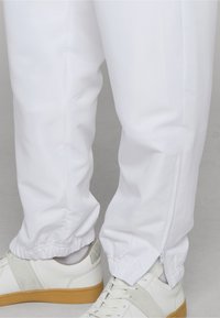 Alsó lábak fehér, elasztikus mandzsettás és oldalsó cipzáros nadrágban, fehér cipővel, barna talppal, szürke padlón.