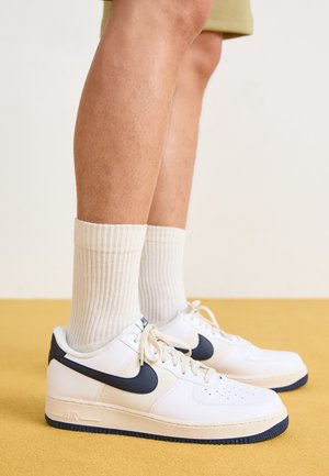 Ben iført beige shorts, hvide ribstrikkede crew-sokker og hvide Nike-sneakers med sort swoosh på gulv.