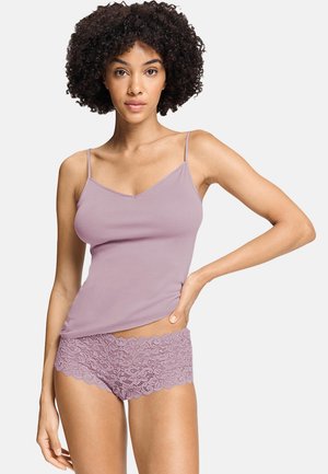 Femme aux cheveux bouclés portant un caraco mauve et un shorty en dentelle assorti, posant avec une main sur la hanche.