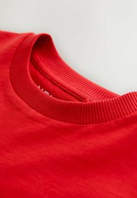 Punase puuvillase t-särgi krae lähedalt vaadatuna, millel on sooniline kaelus ja sile tekstuur. Krae all nähtav märgis.