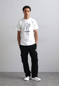 T-shirt blanc avec un graphique d'une cafetière moka et le texte "Moschino", associé à un pantalon cargo noir et des baskets blanches.