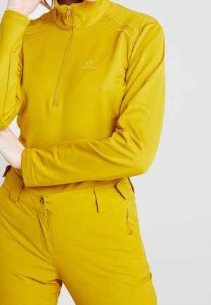 Vrouw draagt mosterdgele lange-mouwen top met rits en bijpassende broek, staand met één hand bij het gezicht en de andere over het lichaam.