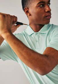 Ljus mintgrön pikétröja med strukturerat tyg, två knappar vid kragen och korta ärmar. Modellen håller i en golfklubba.