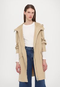 Giacca beige con cappuccio realizzata in tessuto liscio, dotata di zip frontale, tasche laterali e maniche ampie. Indossata sopra una maglietta bianca e jeans blu.