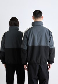 Dos personas llevan chaquetas de colores bloqueados. Las chaquetas tienen un cuello alto, en diferentes tonos de gris, negro y crema, con puños elásticos.