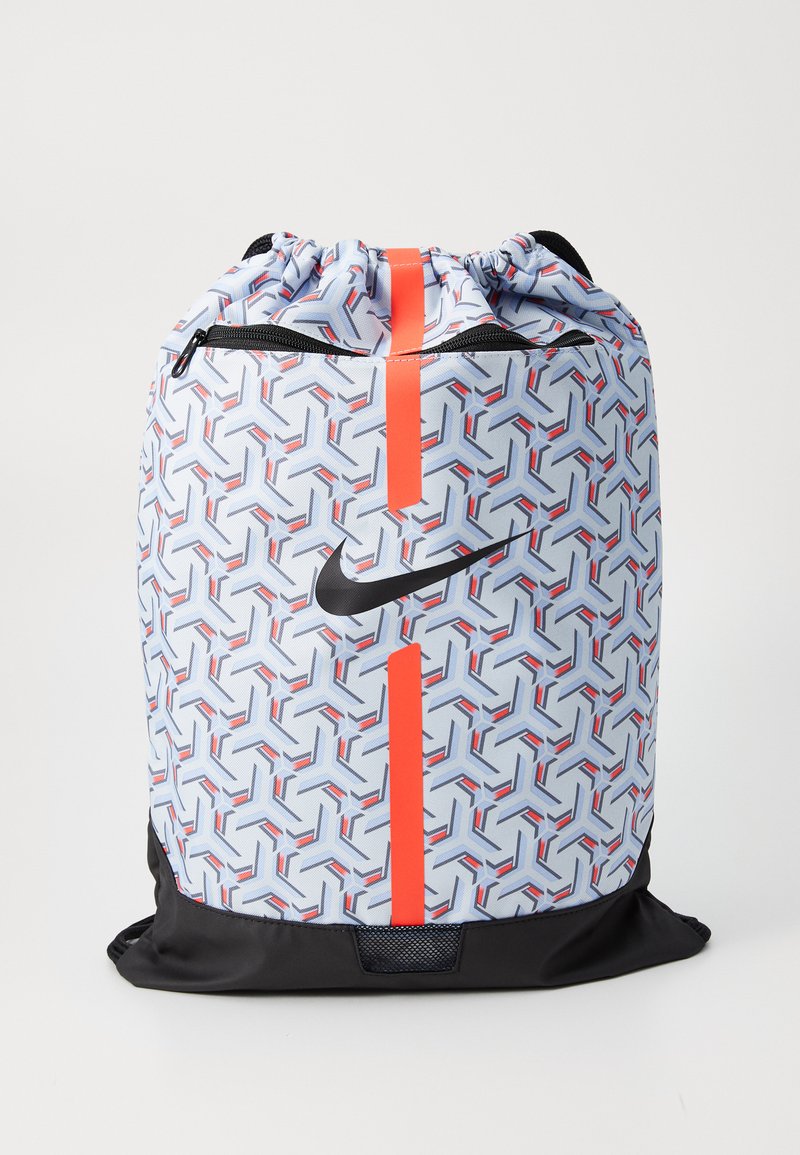 Mochila de cuerda en azul claro con un patrón geométrico en rojo y negro, que presenta un logo de Nike en negro y un bolsillo con cremallera. Base negra.