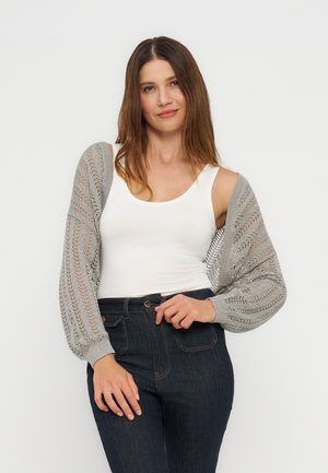 Kobieta o długich brązowych włosach w białym topie na ramiączkach, szarym, luźnym swetrze z balonowymi rękawami oraz ciemnoniebieskich dżinsach z wysokim stanem na białym tle.