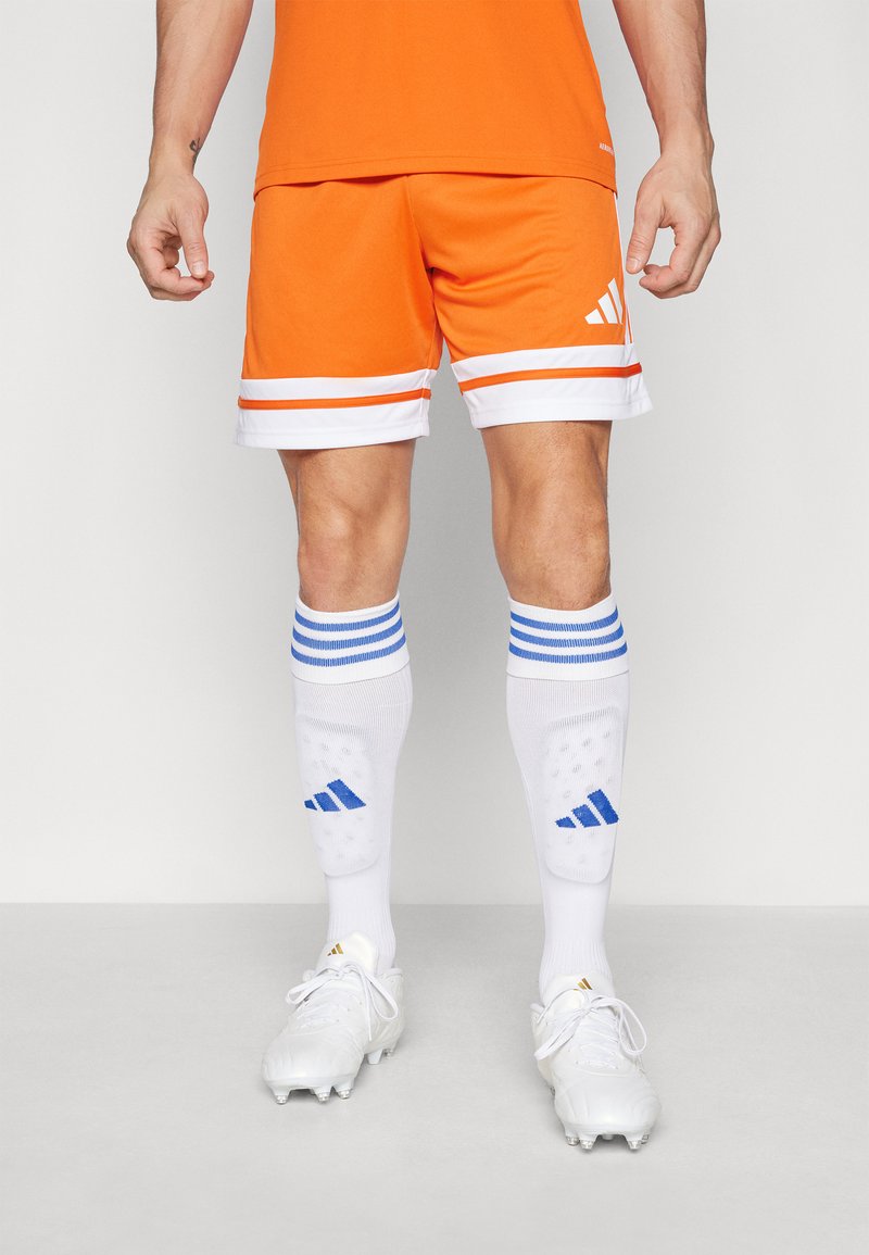 Pantalones cortos deportivos naranjas con rayas blancas, combinados con calcetines blancos con acentos azules y un logo, usados con zapatillas blancas.