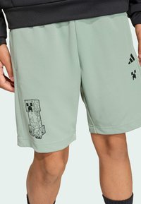 Mätovo zelené športové šortky vyrobené z ľahkého materiálu, s čiernou grafikou Creeper a logom Adidas na prednej strane.