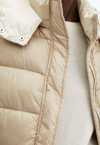 Beige Daunenjacke mit glänzender Oberfläche, versehen mit einem Reißverschluss und einem hohen Kragen. Horizontales Steppdesign mit glatter Textur.