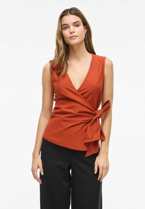 Femme aux longs cheveux portant un haut cache-cœur sans manches couleur rouille avec un nœud sur le côté, associé à un pantalon noir à jambes larges, debout devant un fond blanc.