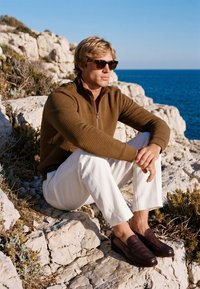 Homme portant des lunettes de soleil, un pull marron, un pantalon blanc et des chaussures foncées assis sur une rive rocheuse avec la mer bleue en arrière-plan.