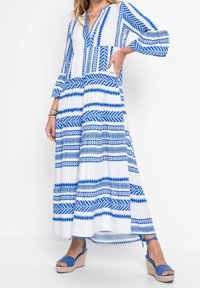 Robe maxi blanche avec des motifs géométriques bleus, présentant un décolleté en V, des manches longues et un bas évasé. Associée à des sandales compensées bleues.