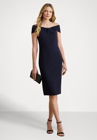 Μπλε σκούρο φόρεμα με ανοιχτούς ώμους και κόμπο στο μπούστο, εφαρμοστή σιλουέτα, μήκος μέχρι το γόνατο, συνδυασμένο με μαύρο τσαντάκι χειρός και πέδιλα με λουράκια.