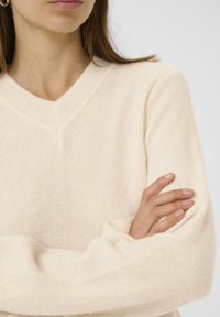 Pull en tricot de couleur crème, doux, avec un décolleté en V, des poignets côtelés et une coupe décontractée. Le tissu a une texture lisse et pelucheuse.