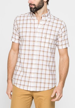 Uomo che indossa una camicia beige e bianca a quadri con maniche corte e pantaloni marrone chiaro, rivolto leggermente a sinistra su uno sfondo semplice.