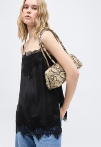 Νεαρή γυναίκα με ξανθά μαλλιά που φοράει μαύρο καλοκαιρινό τοπ με δαντέλα και μπλε τζιν, κρατώντας μια μπεζ τσάντα ώμου με σχέδιο φολιδών φιδιού.