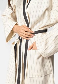 Krem luksuzni ogrtač s crnim pojasom, širokim rukavima i dva prednja džepa. Tkanina izgleda mekano s teksturiranim završetkom.