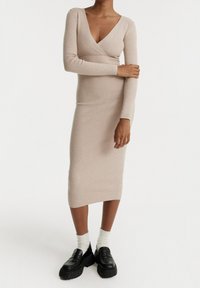 Abito a maglia beige a coste con scollo a V e maniche lunghe, silhouette aderente, lunghezza al ginocchio, abbinato a mocassini neri chunky e calze bianche.