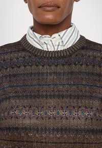 Homme portant une chemise rayée sous un pull en maille à col rond avec des motifs horizontaux multicolores.