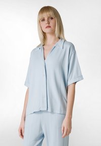 Camisa de manga corta azul claro con cuello, cierre de botones y corte holgado, hecha de una tela suave, con un brillo sutil y un diseño relajado.