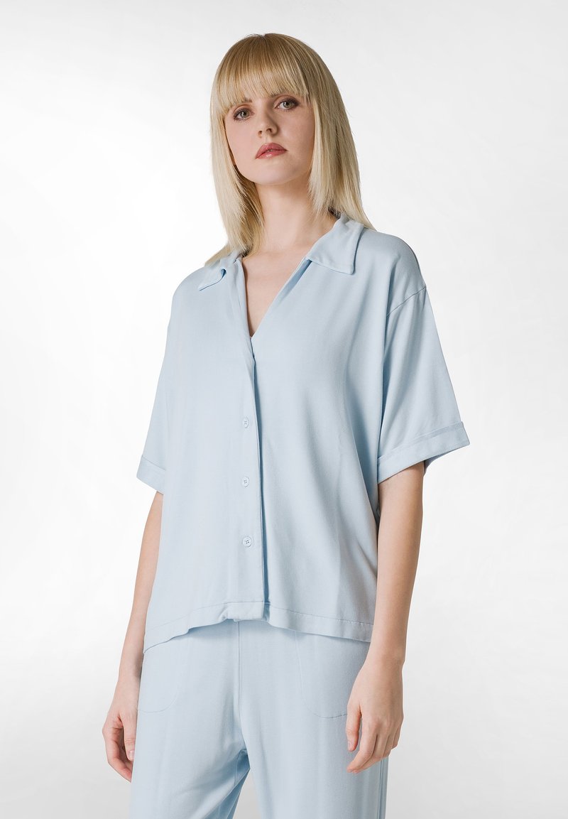 Camisa de manga corta azul claro con cuello, cierre de botones y corte holgado, hecha de una tela suave, con un brillo sutil y un diseño relajado.