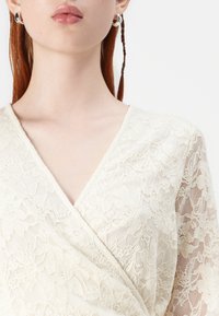 Elfenben blonde topp med dyp V-hals og blomstermønster. Har gjennomsiktige lange ermer og en myk, teksturert overflate.