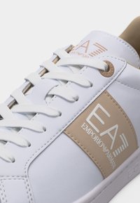 Λευκό αθλητικό παπούτσι Emporio Armani EA7 με μπεζ ετικέτα λογότυπου και λευκά κορδόνια, εμφανιζόμενο σε κοντινή λεπτομέρεια από το πλάι και τη γλώσσα.