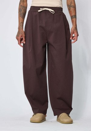 Personne debout portant un pantalon large marron à cordon, des chaussons beiges et un débardeur, les bras visibles avec des tatouages, sur un fond uni.
