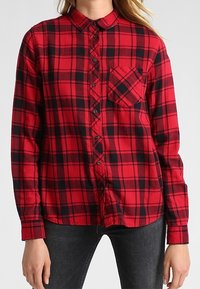 Camisa de franela a cuadros rojos y negros con cuello abotonado, mangas largas y un bolsillo en el pecho. Tejido texturizado con patrón de cuadros.