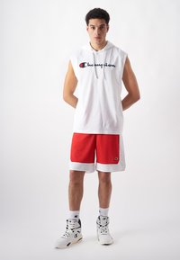 Weißer ärmelloser Hoodie mit "Champion"-Logo, kombiniert mit roten Mesh-Shorts mit weißen Akzenten und weißen Sneakers mit schwarzen Details.
