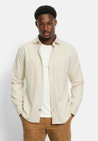 Light beige long-sleeve shirt with buttons and a chest pocket, paired with a white t-shirt and corduroy brown pants. Smooth fabric texture.