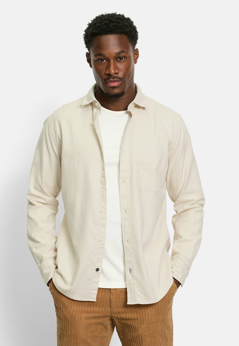Light beige long-sleeve shirt with buttons and a chest pocket, paired with a white t-shirt and corduroy brown pants. Smooth fabric texture.