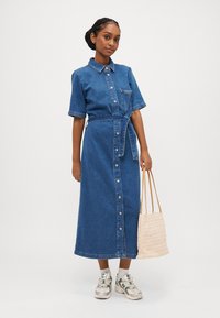 Denim srajčna obleka, kratki rokavi, sprednje gumbno zapiranje, pas z vezavami, dolžina do sredine mečnih. Obuta z pleteno torbico in svetlobo barvnimi športnimi copati.