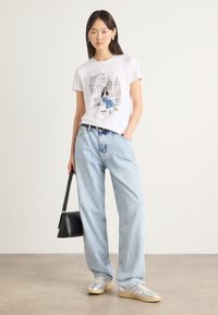T-shirt graphique blanc avec illustration, jean large bleu clair, baskets argentées et sac à main noir. Textures lisses, style décontracté.