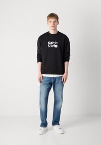 Black sweatshirt with white "Calvin Klein" logo, paired with light blue jeans. Model wears white sneakers, standing against a neutral background.
