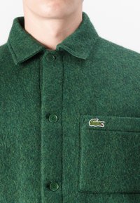 Chemise en laine verte avec un col boutonné, deux boutons à l'avant et une poche poitrine avec un petit logo brodé.