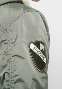Vihreä nailonibomber-takki, jossa on musta-valkoinen merkki, jossa lukee "1st Division" ja tyylitelty emblem. Takki on sileä ja hyvin istuva.