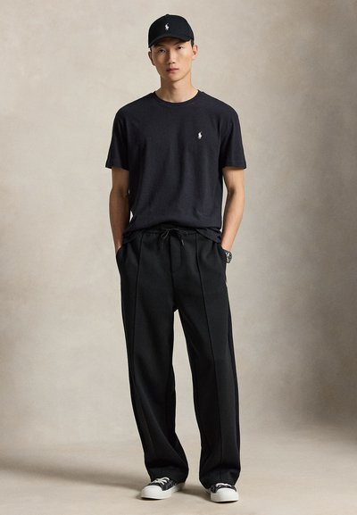 Giovane uomo con berretto nero, t-shirt nera con piccolo logo bianco, pantaloni neri larghi e sneakers nere e bianche, in piedi con le mani in tasca.