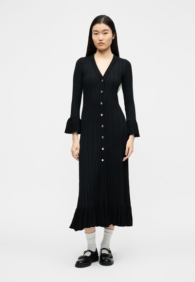 Γυναίκα στέκεται φορώντας μαύρο ριμπ πουκάμισο με κουμπιά, μακρυμάνικο φόρεμα με βολάν στα μανίκια και στο τελείωμα, λευκές κάλτσες και μαύρα μοκασίνια.