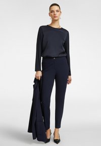 Námořnická hedvábná blůza s dlouhými rukávy, spárovaná s přizpůsobenými námořnickými kalhotami a černými podpatky. Modelka drží sladěný blejzr.