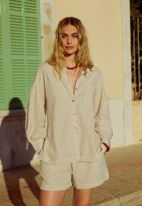 Gestreepte beige button-up shirt met een voork pocket, gecombineerd met bijpassende shorts. Heeft een losse fit en een licht gestructureerde stof. Rode kraalketting.
