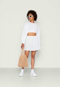 Top cropped branco com olhos de peixe e mangas bufantes, saia rodada combinando, ténis brancos e uma bolsa de palha bege. Design de acessórios minimalista.